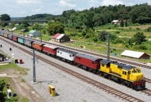 Cena panorâmica de um jogo de construção ferroviária com locomotivas modernas e a vapor, trilhos conectando cidades, pontes, estações movimentadas e paisagem detalhada